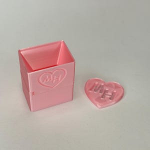 May include: A pink plastic box with a heart-shaped lid. The box and lid have the letters "MHH" embossed on them.