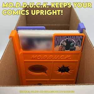 May include: An orange plastic comic book divider with the text MO.D.D.U.C.K. embossed on the front, featuring a rectangular frame for a label. The divider is placed inside a cardboard box to organize comics, with a purple clip on the side.