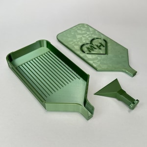 May include: A sage green diamond painting tray with a ribbed interior, a matching lid featuring a heart-shaped emblem with the letters MH, and a small green funnel tool. The set is made of a smooth, matte plastic material with a geometric pattern.
