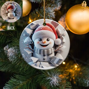 Pode incluir: Um enfeite de Natal redondo de cerâmica branca com um boneco de neve usando um chapéu de Papai Noel vermelho e uma echarpe listrada cinza e branca. O boneco de neve está espiando por um buraco no enfeite, que é decorado com flocos de neve.