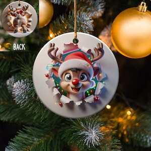 May include: A round white ceramic Christmas bauble featuring a cartoon reindeer wearing a Santa hat peeking through a torn paper hole. The reindeer has a red nose and is surrounded by colourful Christmas lights.