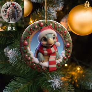 Può includere: Un addobbo natalizio rotondo in ceramica con una tartaruga di cartone animato che indossa un cappello di Babbo Natale rosso e una sciarpa. La tartaruga spunta da una ghirlanda con bacche rosse e foglie verdi.