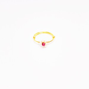May include: A gold-colored wire ring featuring a cluster of small white beads and a central, vibrant red gemstone. The ring is set against a plain white background, highlighting its delicate design and craftsmanship. The ring's band is thin and circular.