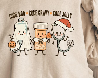 May include: A sand-colored crewneck sweatshirt featuring a graphic of three medical-themed characters with the text CODE BOO, CODE GRAVY, and CODE JOLLY. The characters include an IV bag as a ghost, a pill bottle as a pilgrim, and a stethoscope.