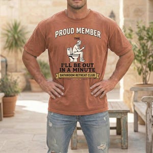 May include: A terracotta-coloured t-shirt with the text "PROUD MEMBER OF THE BATHROOM RETREAT CLUB." The shirt features a graphic of a person on a toilet, looking at their phone. The text "I'LL BE OUT IN A MINUTE" is also printed on the shirt.