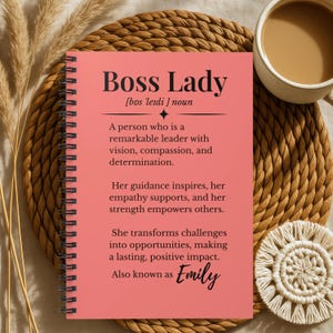 May include: A pink spiral-bound notebook with the words "Boss Lady" and a definition printed on the cover. The notebook is next to a cup of coffee and a decorative coaster. The text also includes the name "Emily".
