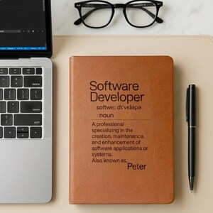 May include: A brown leather notebook with the text "Software Developer" on the cover, including a definition and the name "Peter." A black pen and glasses are also visible on a light-colored surface.