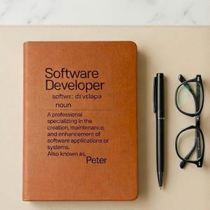 May include: A brown notebook with the text "Software Developer" and a definition. A black pen and black-framed glasses are next to the notebook. The notebook also says "Also known as, Peter."