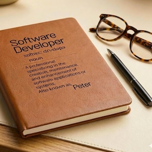 May include: A brown leather notebook with the text "Software Developer" and a definition printed on the cover. Tortoiseshell glasses and a black pen are placed beside the notebook.
