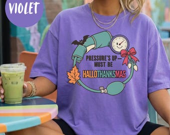 May include: A violet short-sleeve t-shirt featuring a graphic of a blood pressure cuff shaped into a wreath. The design includes a bat, an orange maple leaf, a red bow, and the text: PRESSURE'S UP - MUST BE HALLOTHANKSMAS.
