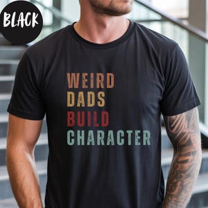May include: Black t-shirt with the text "WEIRD DADS BUILD CHARACTER" in a retro font. The word "BLACK" is in a white circle in the upper left corner. The shirt is a crew neck and has short sleeves.