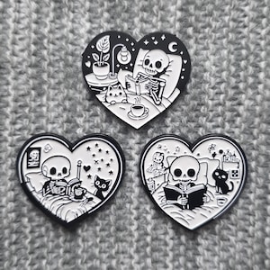 Pode incluir: Três pinos de esmalte preto e branco em forma de coração. Cada pino apresenta um esqueleto lendo um livro na cama com um gato. O primeiro pino tem uma planta e uma xícara de chá. O segundo pino tem um céu estrelado. O terceiro pino tem uma mesa de cabeceira com uma lâmpada.
