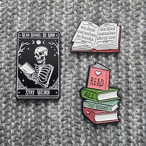 Può includere: Tre spille in smalto con disegni a tema libro. La prima spilla presenta uno scheletro che legge un libro con il testo "Read Books, Be Kind" e "Stay Weird". La seconda spilla presenta un libro aperto con il testo "We lose ourselves in books" e "We find ourselves there too". La terza spilla presenta una pila di libri con il testo "Read", "All", e "The Books".