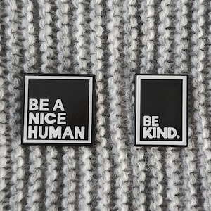 Puede incluir: Dos pines de esmalte negro con letras blancas. El primer pin dice "Be a Nice Human". El segundo pin dice "Be Kind."