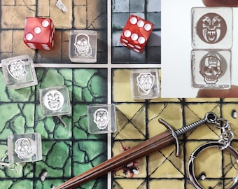Pode incluir: Um conjunto de dados com desenhos de caveiras e escudos, um chaveiro de espada e dois dados vermelhos com pontos brancos. Os dados estão em um tabuleiro de jogo verde e marrom.
