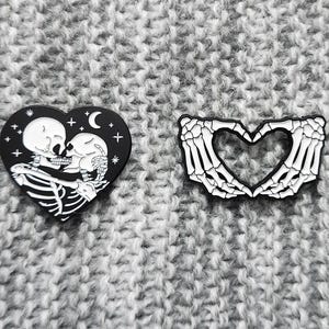 May include: Two black enamel pins with a white skeleton design. One pin features a skeleton couple kissing inside a heart shape with stars and a crescent moon. The other pin features two skeleton hands forming a heart shape.