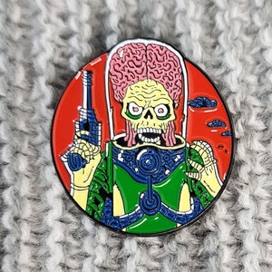 Op de afbeelding: Een rode, witte en blauwe emaille pin met een cartoon-buitenaardse met een blootgesteld brein, die een pistool vasthoudt. De buitenaardse heeft een groen lichaam en een roze brein. De pin is rond en heeft een zwarte omtrek.