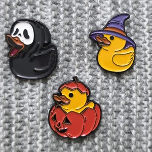 May include: Three Halloween-themed enamel pins featuring duck designs. One is a black duck in a Scream mask, another is a yellow duck wearing a witch hat, and the third is a duck emerging from a carved pumpkin. The pins have a glossy finish.
