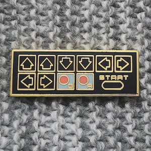 May include: A black and gold enamel pin with a retro video game controller design. The pin features directional arrows, two red buttons, and a "START" button.