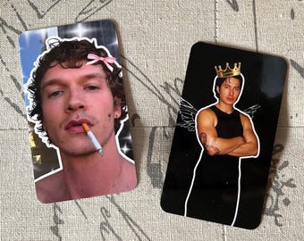 May include: Two rectangular stickers. One features a man with curly hair, a cigarette, and a pink bow. The other shows a man in a black tank top, a gold crown, and angel wings, outlined in white against a black background.