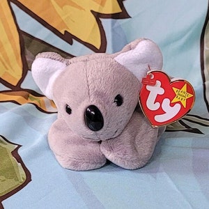 May include: A gray koala beanie baby with a red Ty tag that says "Beanie Babies" and has a star on it. The beanie baby is sitting on a blue and yellow fabric background.