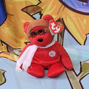 May include: Red beanie baby bear wearing aviator sunglasses and a white scarf. The bear is sitting on a blue and yellow fabric with a peace sign and sunflowers.