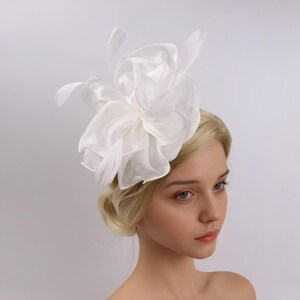 Pode incluir: Acess&oacute;rio de cabelo branco com pena e flor de tule. A flor &eacute; grande e em camadas, com uma textura delicada e enrugada. As penas s&atilde;o macias e esvoa&ccedil;antes, adicionando um toque de fantasia ao design.