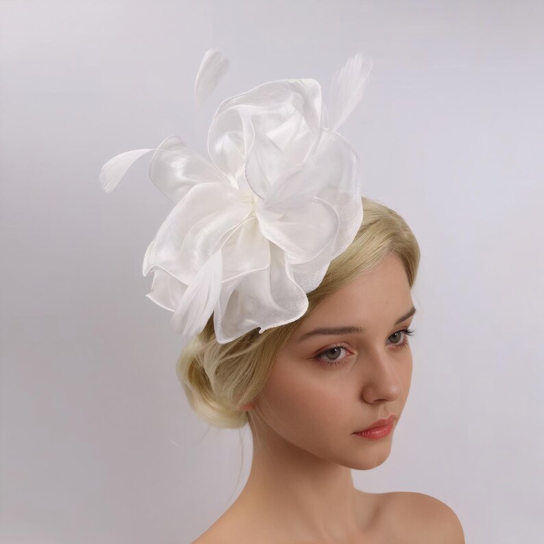 Pode incluir: Acess&oacute;rio de cabelo branco com pena e flor de tule. A flor &eacute; grande e em camadas, com uma textura delicada e enrugada. As penas s&atilde;o macias e esvoa&ccedil;antes, adicionando um toque de fantasia ao design.