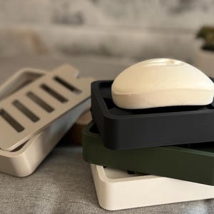 May include: A stack of three rectangular soap dishes in black, olive green, and white, each featuring a removable slotted tray. A single smooth, off-white oval bar of soap rests on the top black dish, while a separate beige soap dish sits nearby.