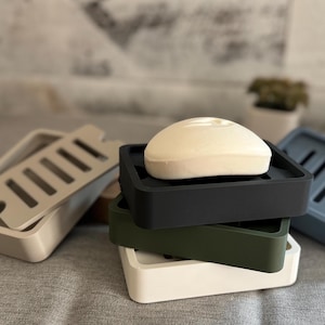 May include: A stack of soap dishes in various colors: off-white, black, olive green, and blue. Each dish has a rectangular shape with a slotted design for drainage. A bar of white soap rests on the top black dish.