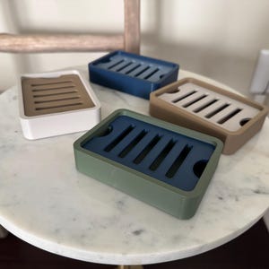 May include: Four rectangular soap dishes in different colours: white with tan insert, blue, tan with white insert, and green with blue insert. Each dish has a slotted design for drainage. The dishes are displayed on a white marble table.