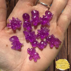 May include: A collection of purple glitter dinosaur charms. The charms are made of resin and have a sparkly finish. They are perfect for adding a touch of whimsy to jewellery, crafts, and other projects.