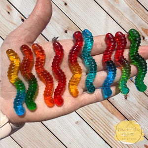 May include: A hand holding a set of eight colorful gummy worm charms. The charms are made of clear plastic and are shaped like gummy worms. They are in a rainbow of colors, including red, orange, yellow, green, blue, and purple.