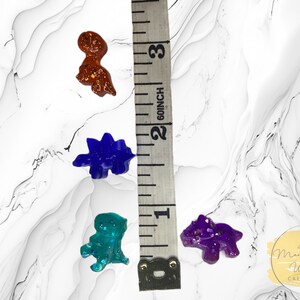 May include: Four small dinosaur figurines made of resin with glitter embedded in the clear resin. The dinosaurs are in shades of purple, blue, green, and brown. The dinosaurs are approximately 2.5 cm in size.