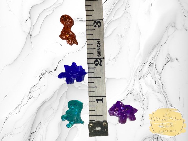 May include: Four small dinosaur figurines made of resin with glitter embedded in the clear resin. The dinosaurs are in shades of purple, blue, green, and brown. The dinosaurs are approximately 2.5 cm in size.