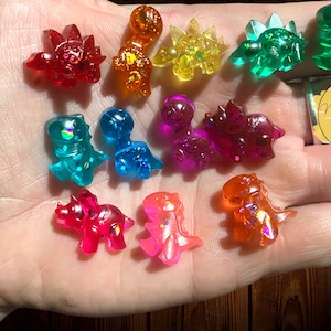 May include: A collection of 10 small, colorful dinosaur figurines made of resin. The dinosaurs are all different colors, including red, green, blue, yellow, orange, and purple. They are all about 1 inch tall and have glitter embedded in the resin.