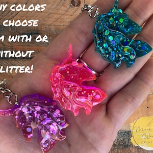 May include: Three unicorn keychains in pink, purple, and teal with glitter. The keychains are made of resin and have a silver chain.