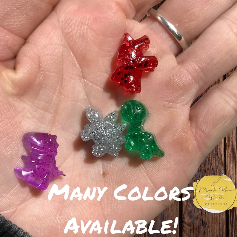 May include: Five small, glittery resin figurines in various colours, including red, green, silver, and purple. The figurines are shaped like a unicorn, a dinosaur, a turtle, and two other dinosaurs. The text "MANY COLORS AVAILABLE!" is written below the figurines.
