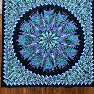 May include: A quilt with a large, teal and purple starburst design on a black background. The starburst is surrounded by a teal and purple border.