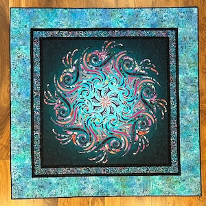 May include: A teal blue quilted fabric square with a black frame and a colorful mandala design in the center. The mandala features swirling patterns in shades of pink, blue, green, and purple.
