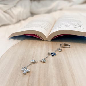 Puede incluir: Una pulsera de plata con varios dijes, incluyendo un corazón, una estrella y un trébol de cuatro hojas. La pulsera se muestra sobre una superficie de madera junto a un libro abierto.