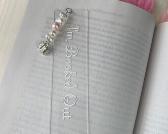 Puede incluir: Un marcapáginas de acrílico transparente con el texto "I'm Booked Out" en letras blancas. El marcapáginas está decorado con un dije plateado en forma de libro y una cadena de perlas blancas y rosadas.
