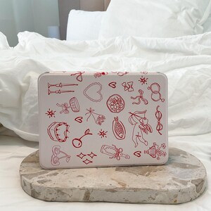 May include: White metal tin with a red and white design. The tin is decorated with various illustrations of hearts, cherries, stars, bows, and other romantic symbols.