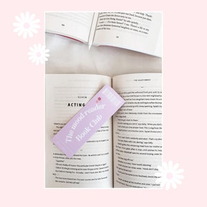 Könnte beinhalten: Ein violettes Lesezeichen mit dem Text "The mood reader Book Club" in weißen Buchstaben. Das Lesezeichen ist mit einer kleinen weißen Blume und einem weißen Stern verziert. Das Lesezeichen liegt offen auf einem Buch mit dem sichtbaren Text "The Glass House".