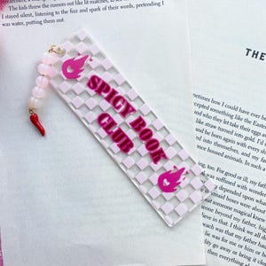 May include: A pink and white bookmark with a checkered pattern and the words "Spicy Book Club" in pink lettering. The bookmark has a pink flame design with a heart at the top.