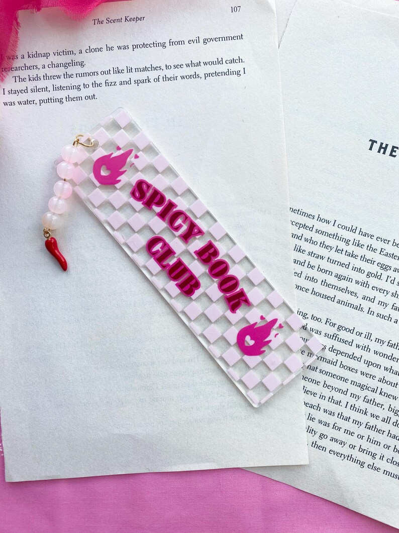 May include: A pink and white bookmark with a checkered pattern and the words "Spicy Book Club" in pink lettering. The bookmark has a pink flame design with a heart at the top.
