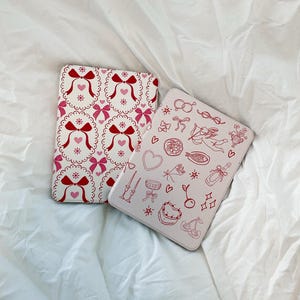 May include: Two pink and white patterned iPad cases. One case has a repeating pattern of red bows and hearts. The other case has a pink and white pattern of hearts, bows, cherries, stars, and other small details.