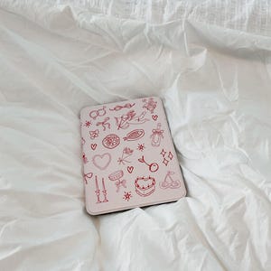 May include: A pink laptop case with a red design of various hearts, cherries, stars, and other symbols. The case is on a white bedspread.