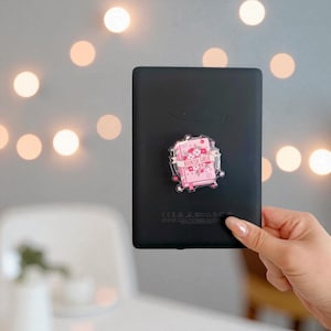 May include: Black e-reader with a decorative pop socket. The pop socket features a pink book design with the words "BOOKISH GIRLIE" and floral accents. The background has soft, blurred lights.