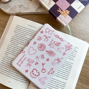 May include: A white notebook cover with a red and white design featuring cherries, stars, bows, and other small illustrations. The cover has a subtle sparkle finish.